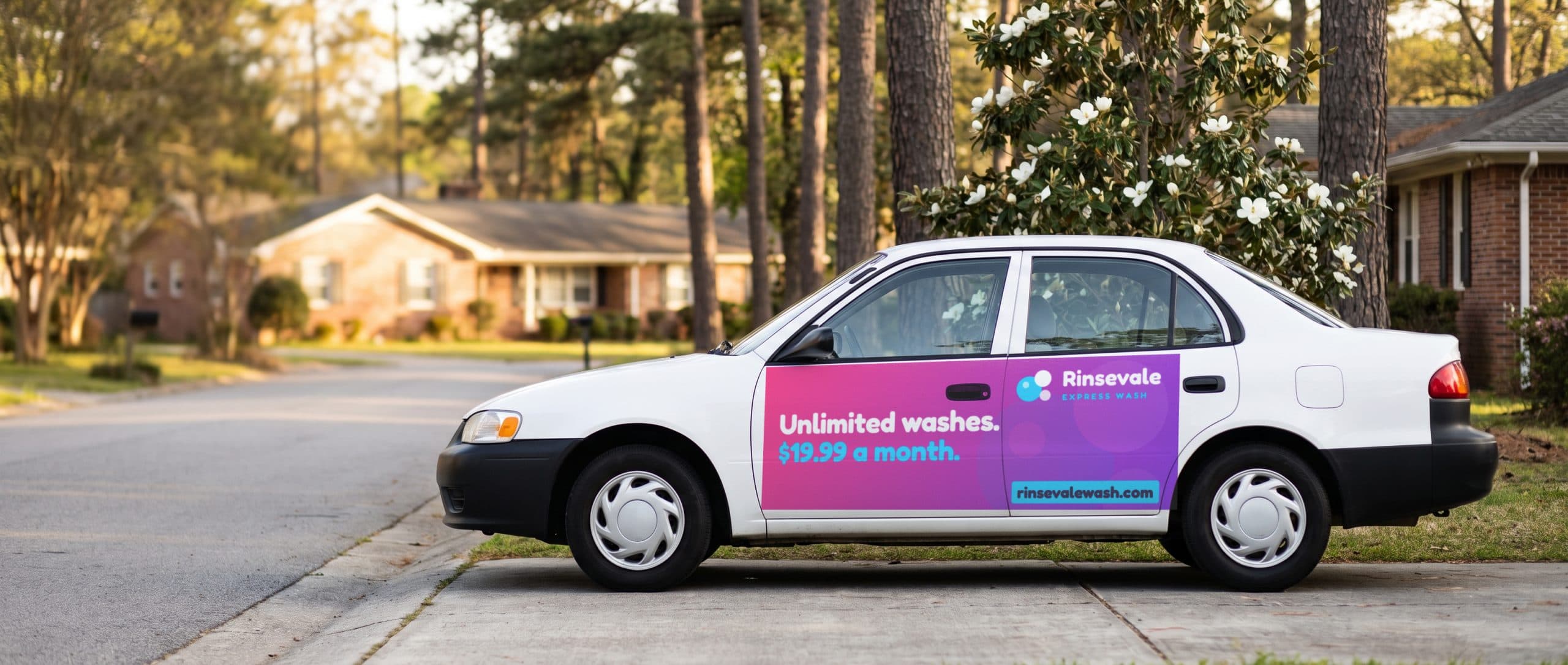 A white sedan parked on a tree-lined suburban street with a car wash ad wrapped across its doors.